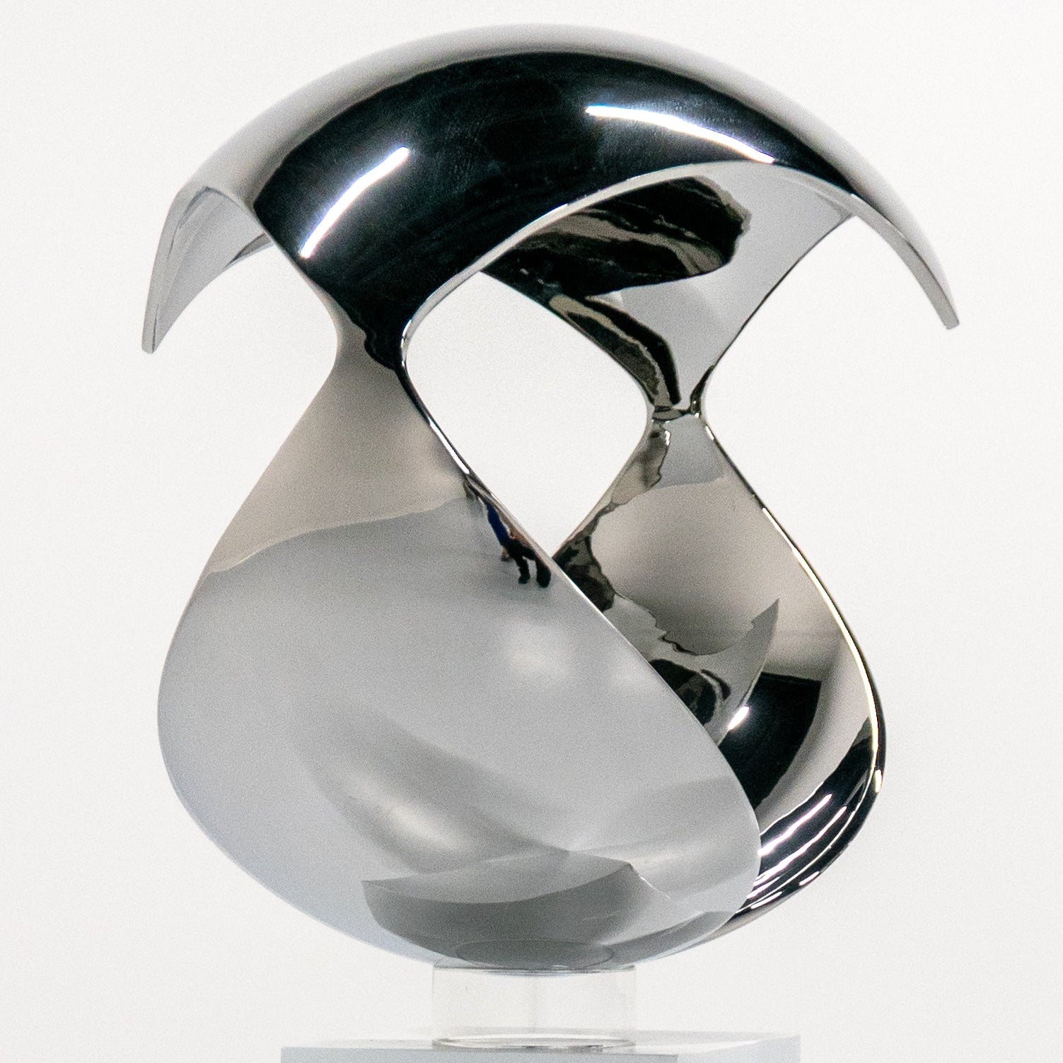A polished chrome abstract sculpture mounted on a white rectangular pedestal, photographed in a clean white studio setting.